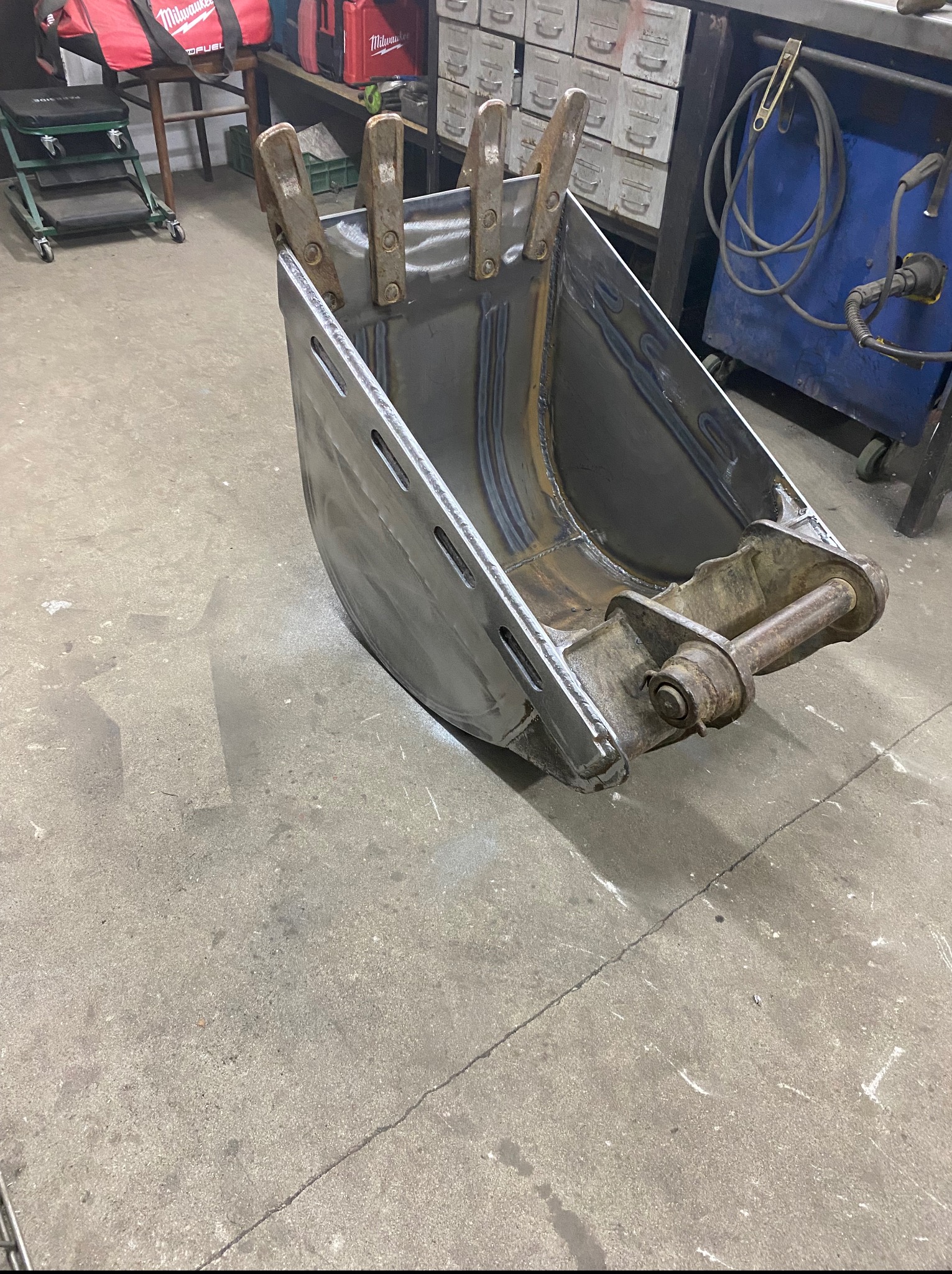 Szara, metalowa łyżka koparki z uchwytami, widoczne spawy, na betonowej posadzce warsztatu, z narzędziami w tle. Detal wykonania i solidność konstrukcji.