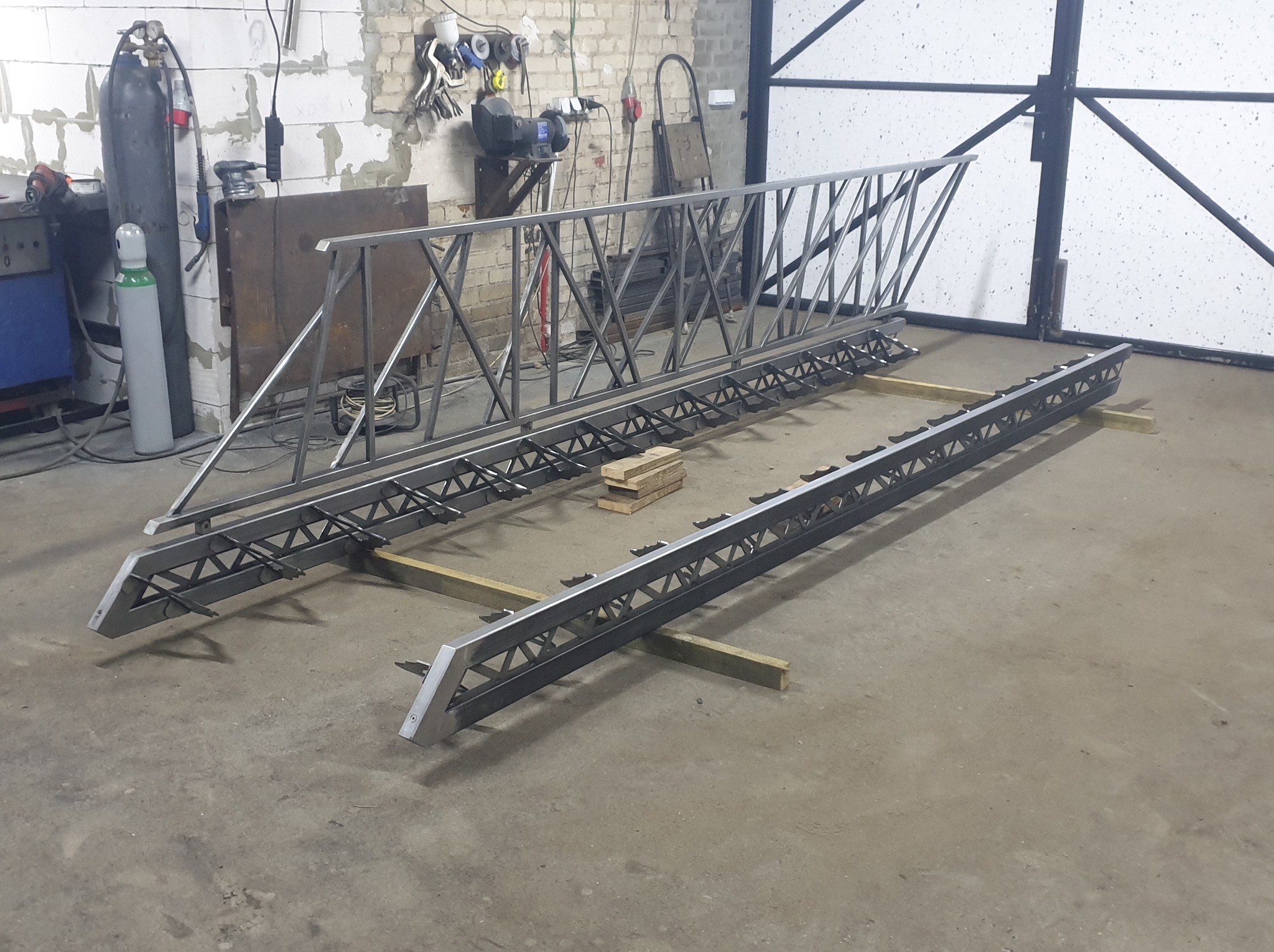 Surowe, metalowe elementy konstrukcyjne z kratownicą, prawdopodobnie fragmenty balustrady lub ogrodzenia, w warsztacie. Widoczne narzędzia i butle spawalnicze w tle.