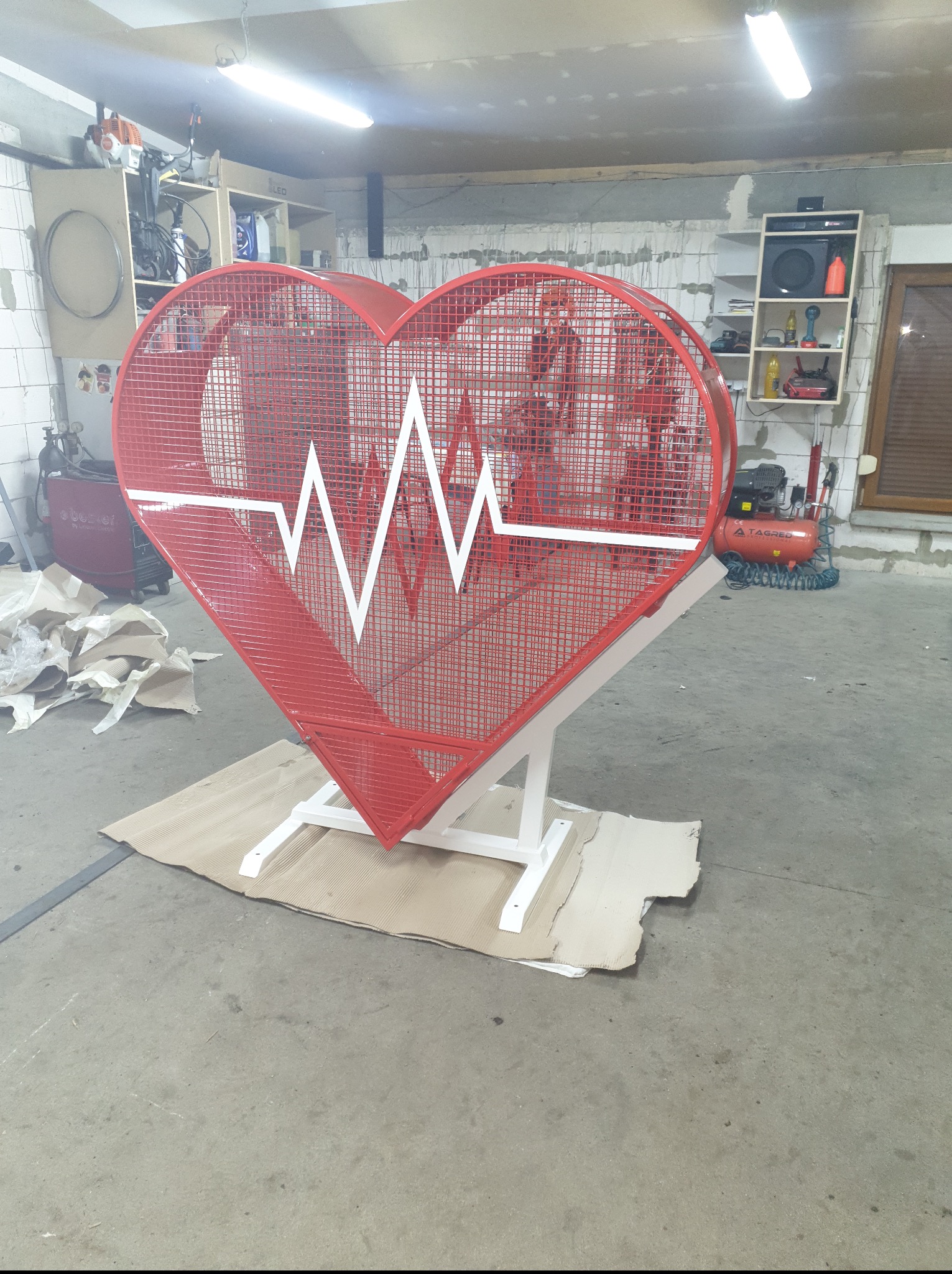 Czerwona, metalowa konstrukcja w kształcie serca z białym motywem EKG, umieszczona na białym stelażu. Wykonana z siatki. Stoi w warsztacie na kartonie.