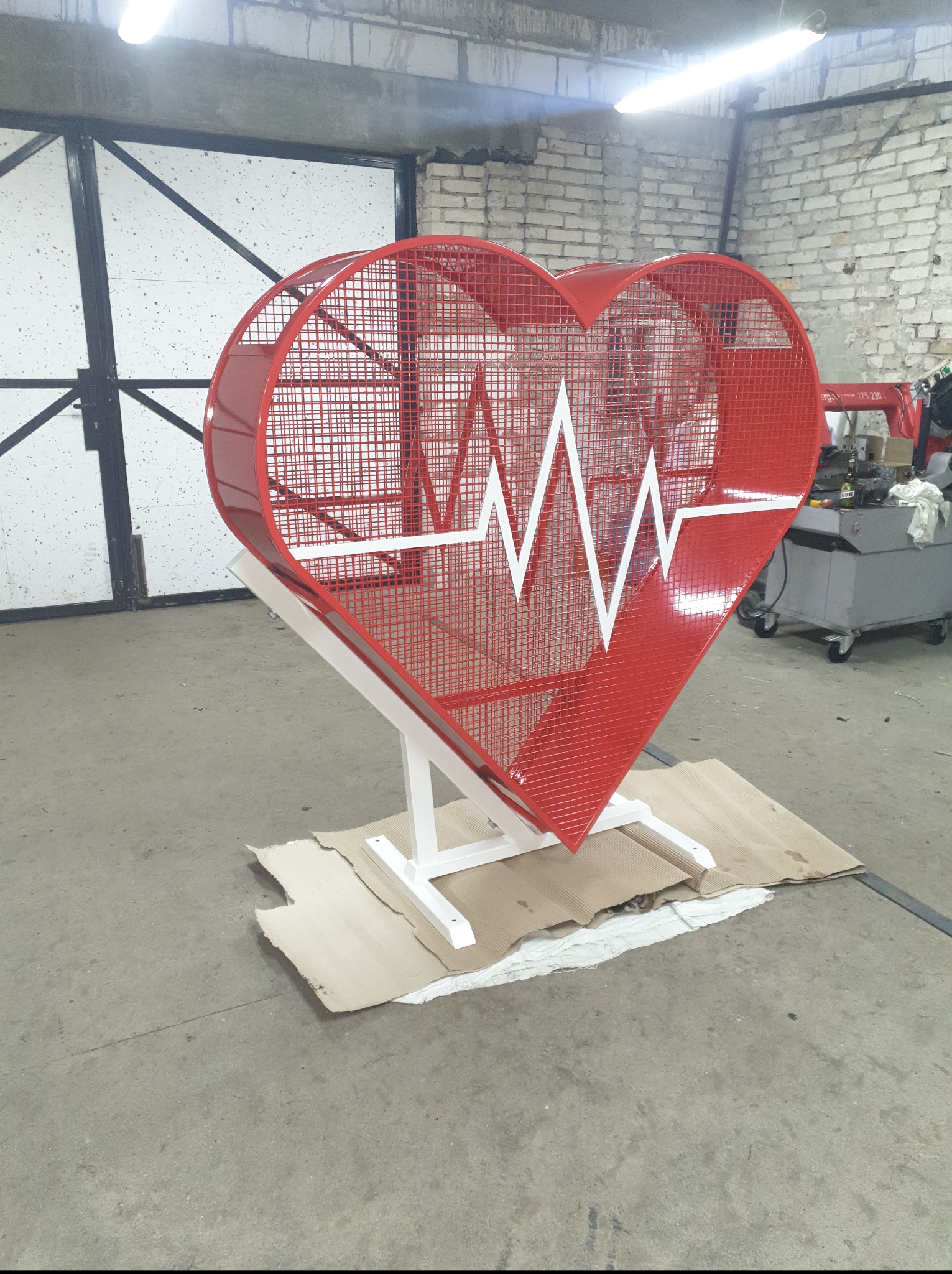 Czerwona, metalowa konstrukcja w kształcie serca z białym symbolem EKG, umieszczona na białym stelażu w warsztacie. Nietypowa dekoracja.