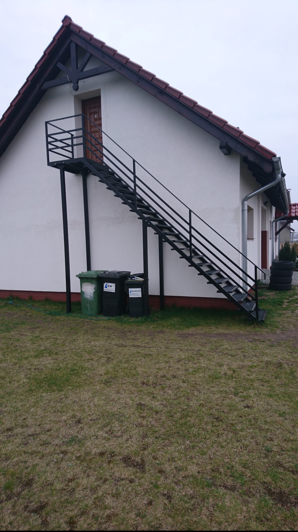Zewnętrzne schody metalowe z czarną, prostą balustradą prowadzące do drzwi na piętrze budynku. Na pierwszym planie widoczne pojemniki na śmieci i trawnik.