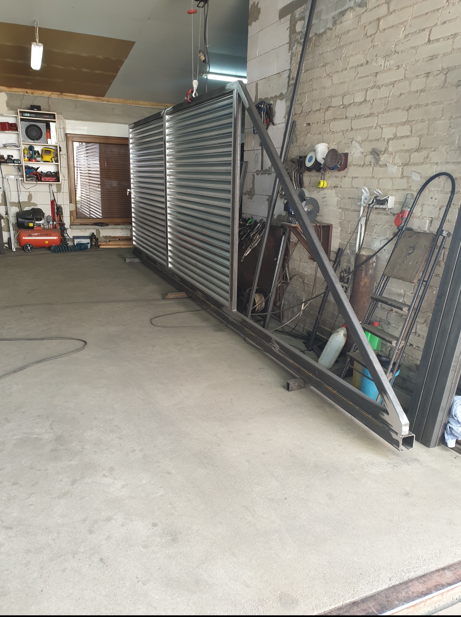 Warsztatowa fotografia ukończonej, metalowej konstrukcji bramy z wypełnieniem z blachy trapezowej, oparta o ścianę z cegły. Widoczne narzędzia i akcesoria ślusarskie.