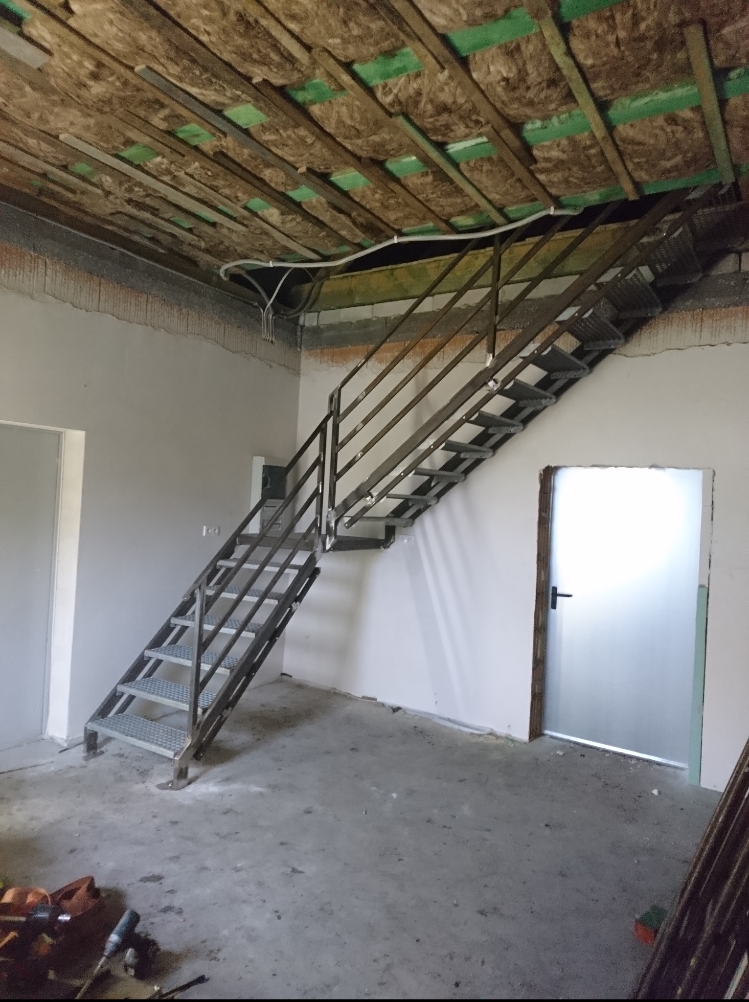 Metalowe schody z ażurowymi stopniami w surowym wnętrzu. Widoczne ocieplenie stropu i narzędzia na podłodze. Ujęcie z dołu, podkreślające konstrukcję.