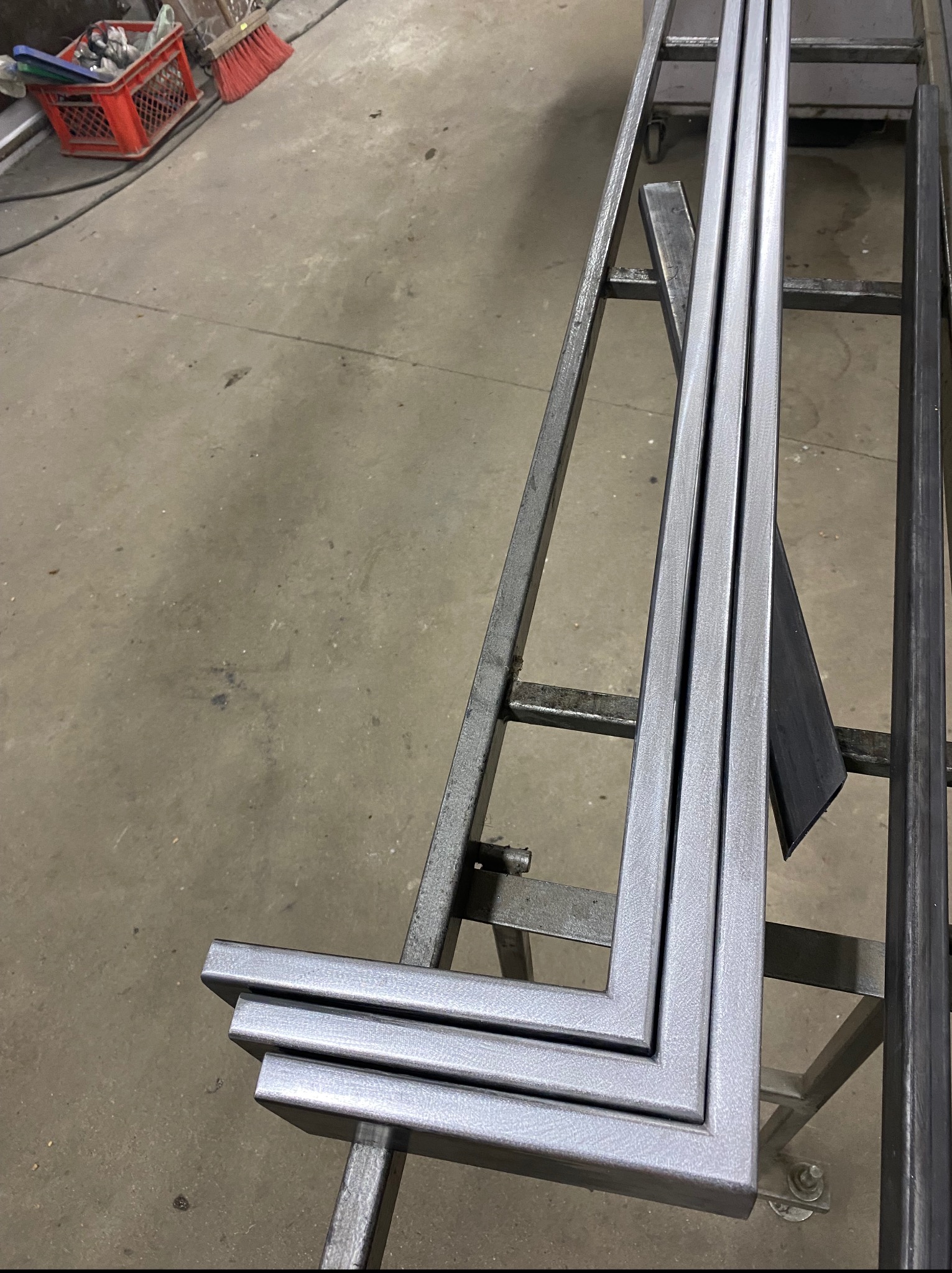 Kątowniki ze stali, trzy srebrne profile ułożone na metalowym stojaku w warsztacie. Widoczne ślady obróbki i surowy beton na podłodze.