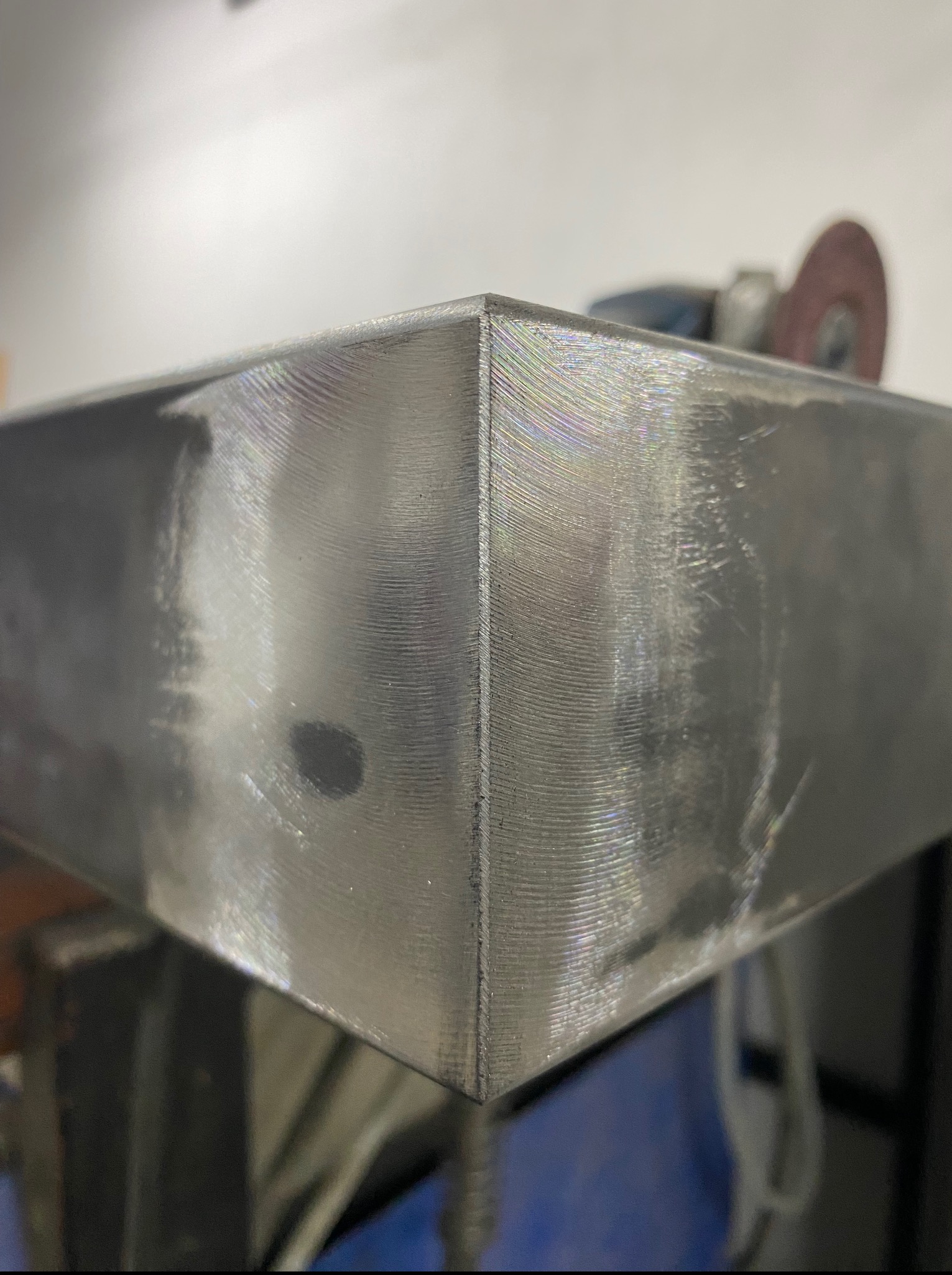 Szlifowany, metalowy narożnik z widocznymi śladami obróbki i tęczowymi refleksami światła. W tle szlifierka kątowa.