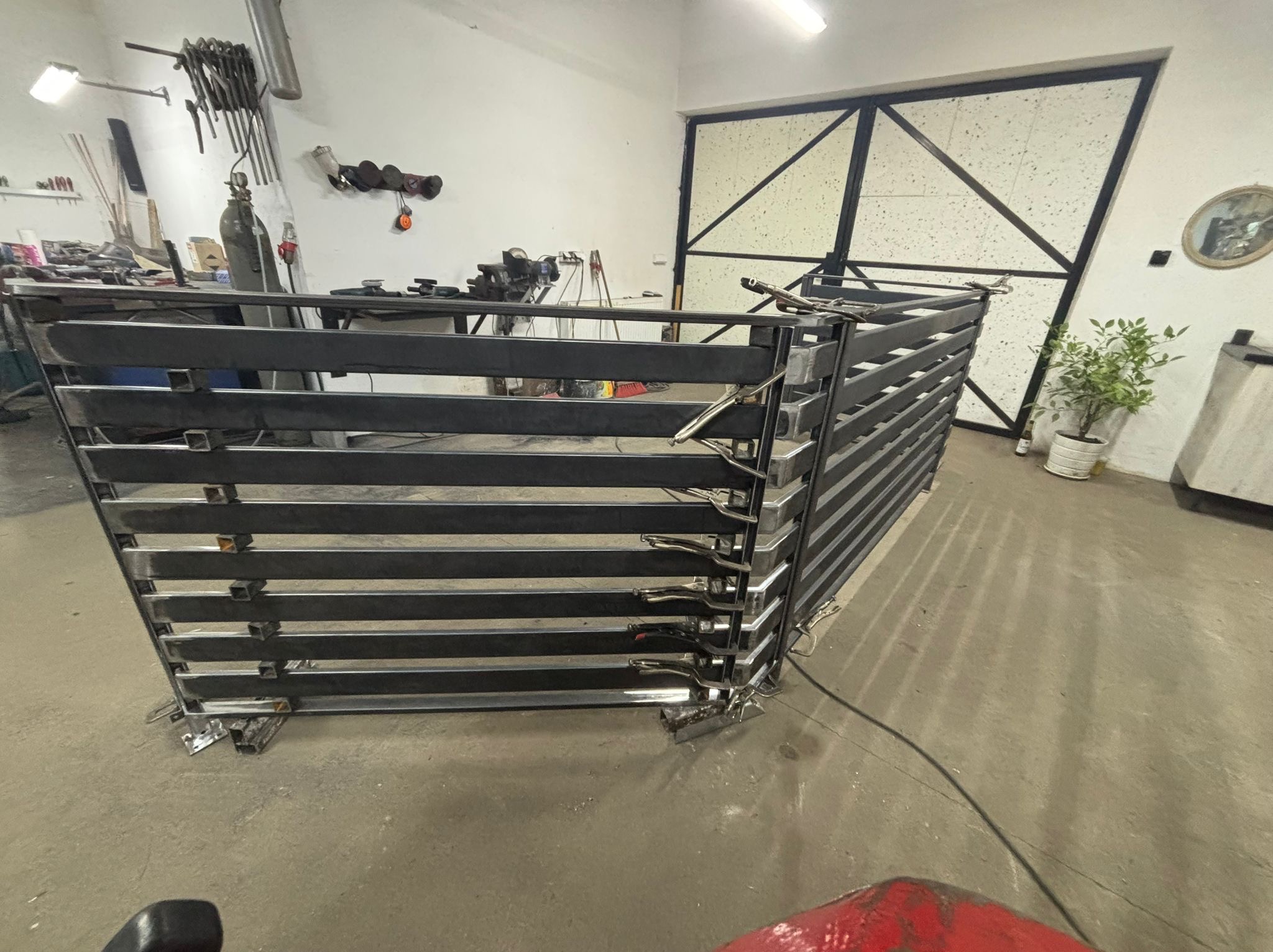 Warsztatowe ujęcie metalowej konstrukcji z poziomych profili, zabezpieczonej klamrami spawalniczymi, na tle białej ściany z narzędziami i metalowej bramy.