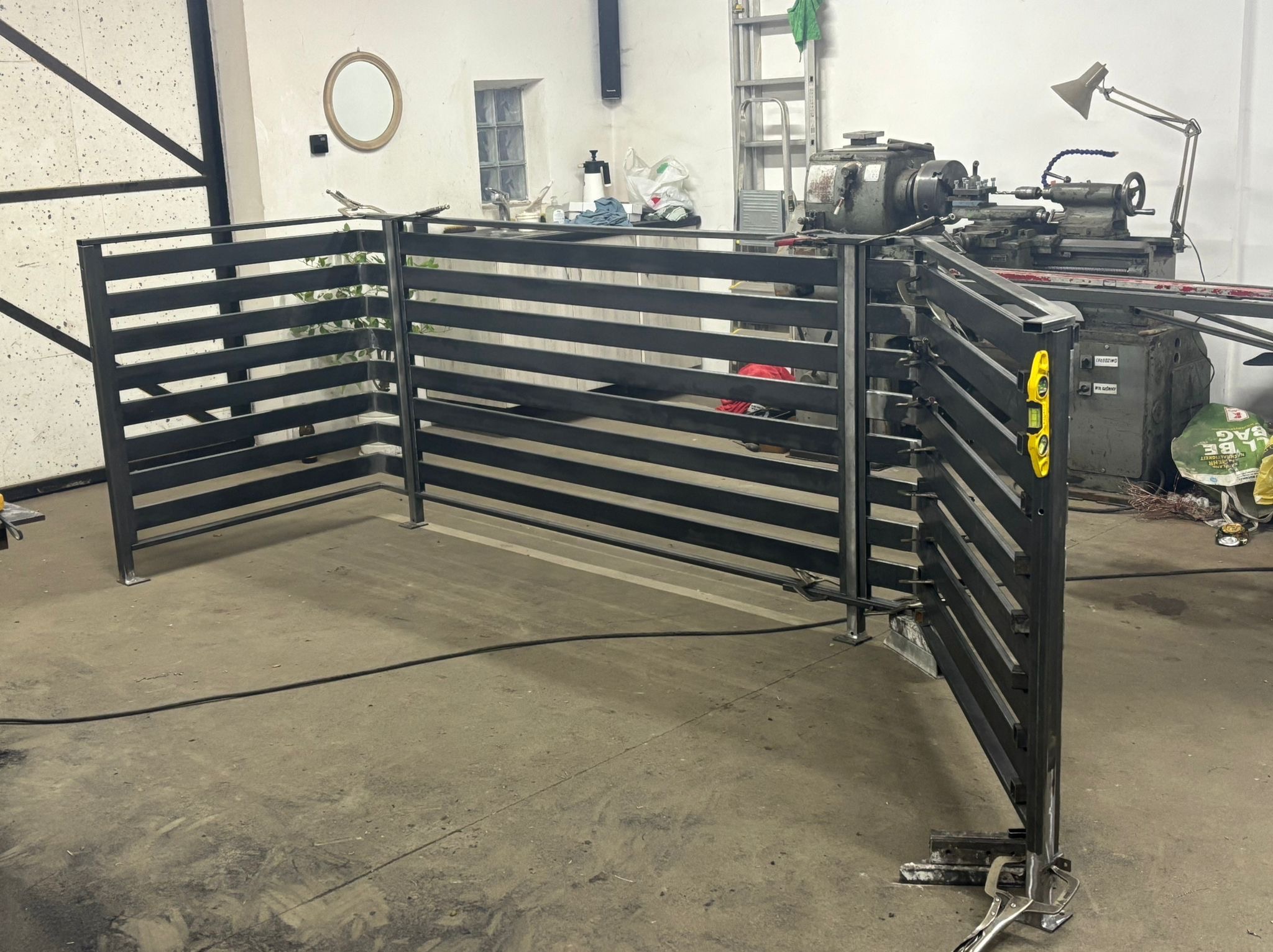 Częściowo zmontowane, czarne, metalowe ogrodzenie panelowe w warsztacie. Widoczny poziomica, spawarka i narzędzia ślusarskie. Surowe wykończenie podłogi.