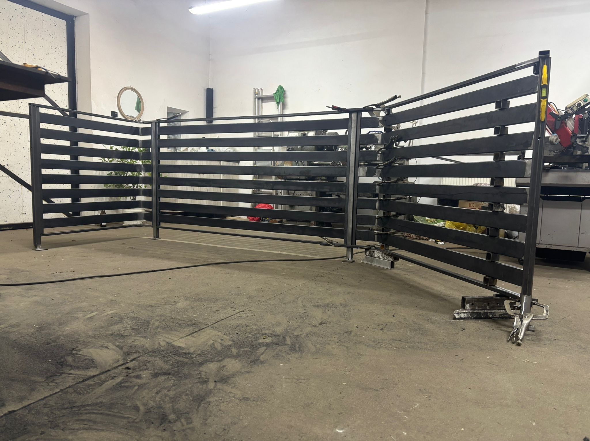 Metalowa konstrukcja ogrodzenia w warsztacie, widoczne narzędzia i elementy spawania. Surowy stan, przed malowaniem. Oświetlenie warsztatowe.