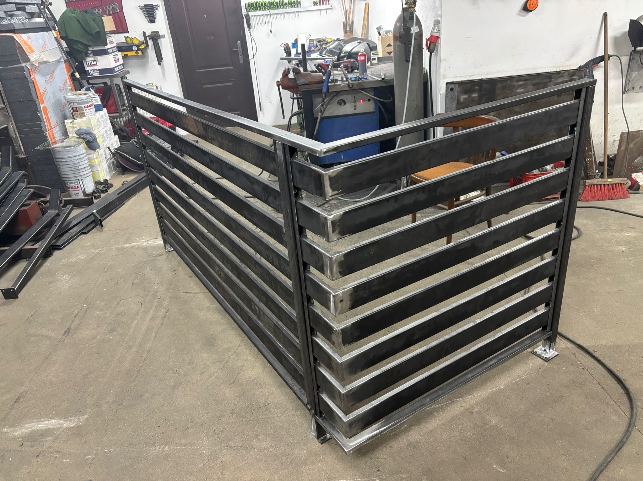 Surowa, metalowa konstrukcja balustrady z poziomymi szczeblami, spawana w warsztacie, widoczne ślady obróbki, surowy beton na podłodze, narzędzia w tle.