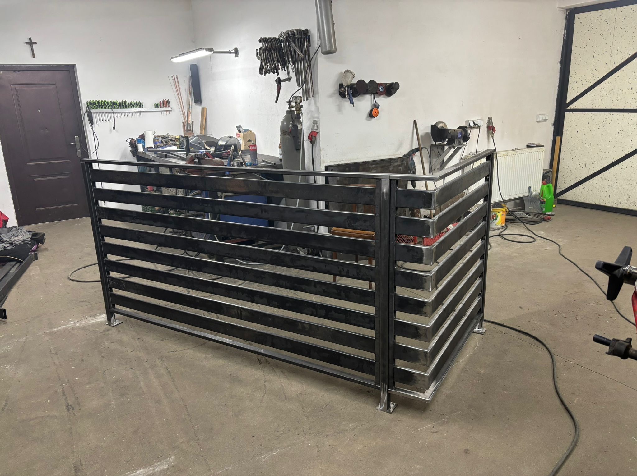 Surowa, metalowa konstrukcja z poziomych profili w warsztacie. Widoczne narzędzia i sprzęt spawalniczy w tle. Element balustrady lub ogrodzenia w trakcie produkcji.