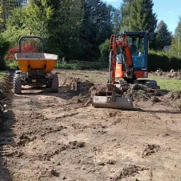 Ż&oacute;łta wywrotka i pomarańczowa koparka na placu budowy z wykopaną ziemią, w tle drzewa i niebieskie niebo. Prace ziemne w toku.