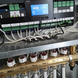 Instal Adam Mróz - Rozdzielacz ogrzewania podłogowego z widocznym sterownikiem, przewodami i opisami stref grzewczych. Instalacja w metalowej obudowie, widok z bliska.