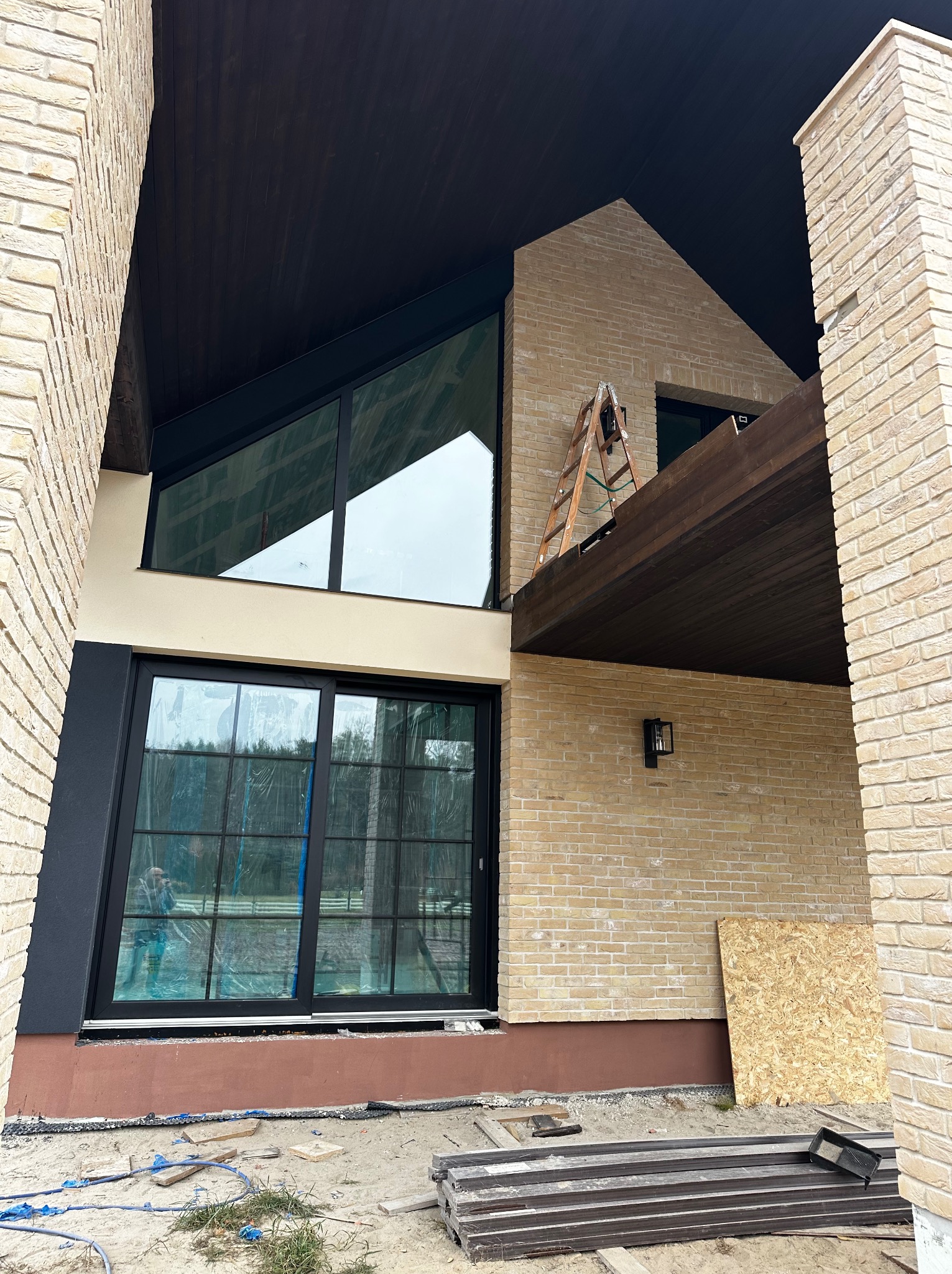 Elewacja domu z jasnej cegły klinkierowej, duże okna z czarnymi ramami, drewniane podbitki. Na zewnątrz widoczne materiały budowlane i drabina.