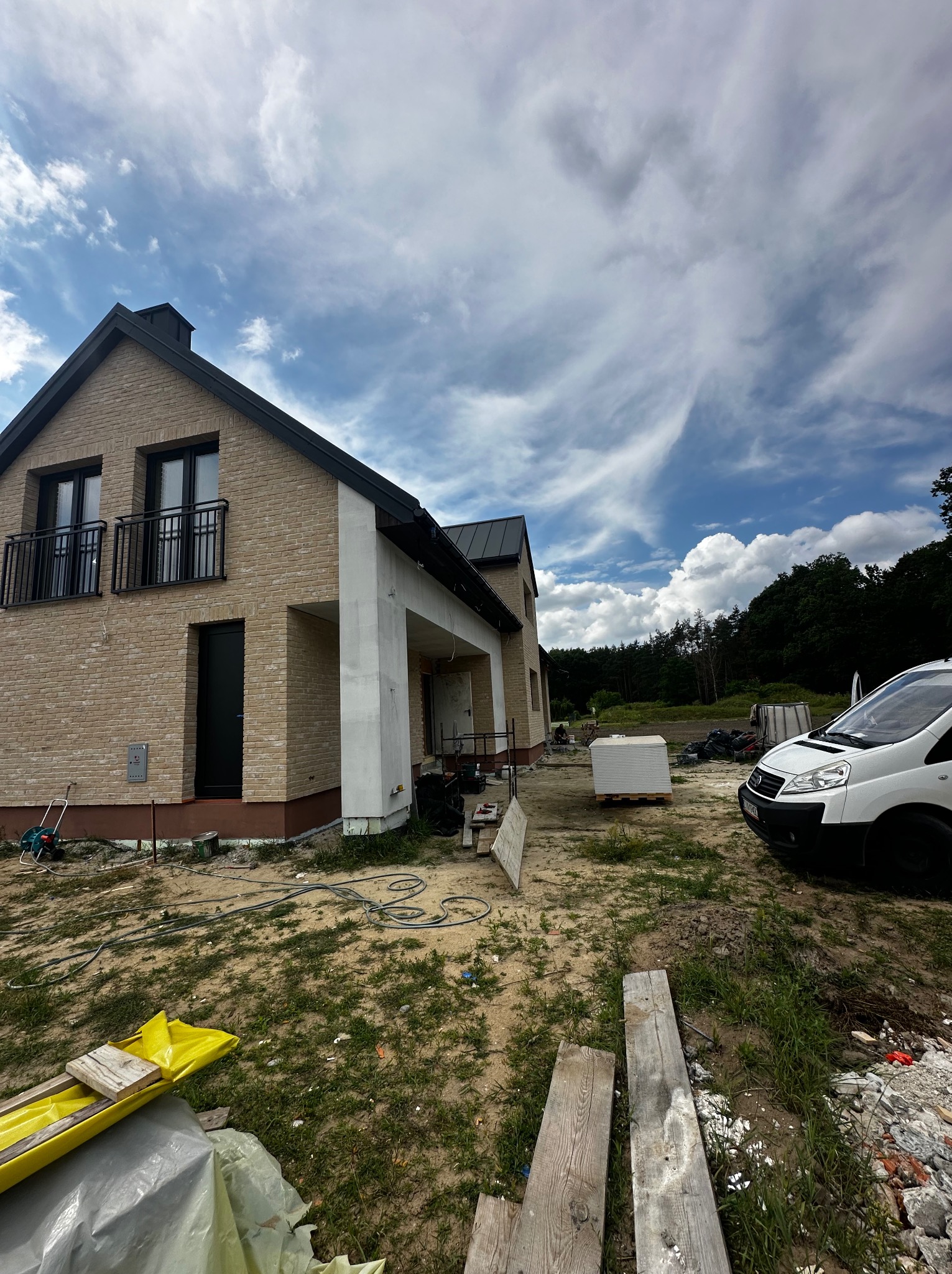 Nowoczesny dom z cegły klinkierowej w trakcie budowy, widok z zewnątrz, z elementami wykończeniowymi i materiałami budowlanymi na posesji. Biały samochód dostawczy w tle.