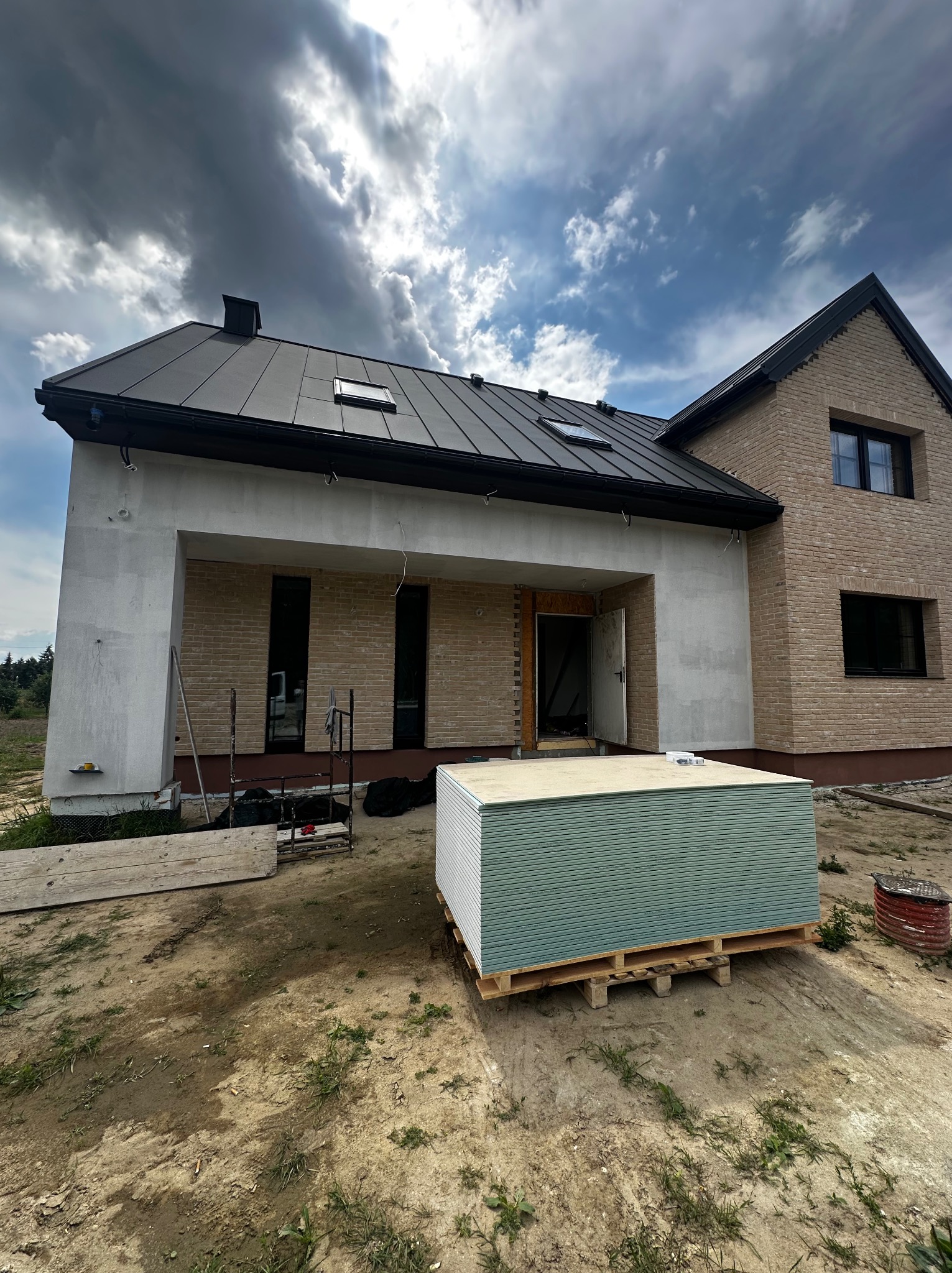 Dom w trakcie budowy z widocznymi płytami na palecie. Część budynku wykończona cegłą, druga w stanie surowym. Na dachu okna dachowe. Pochmurne niebo.