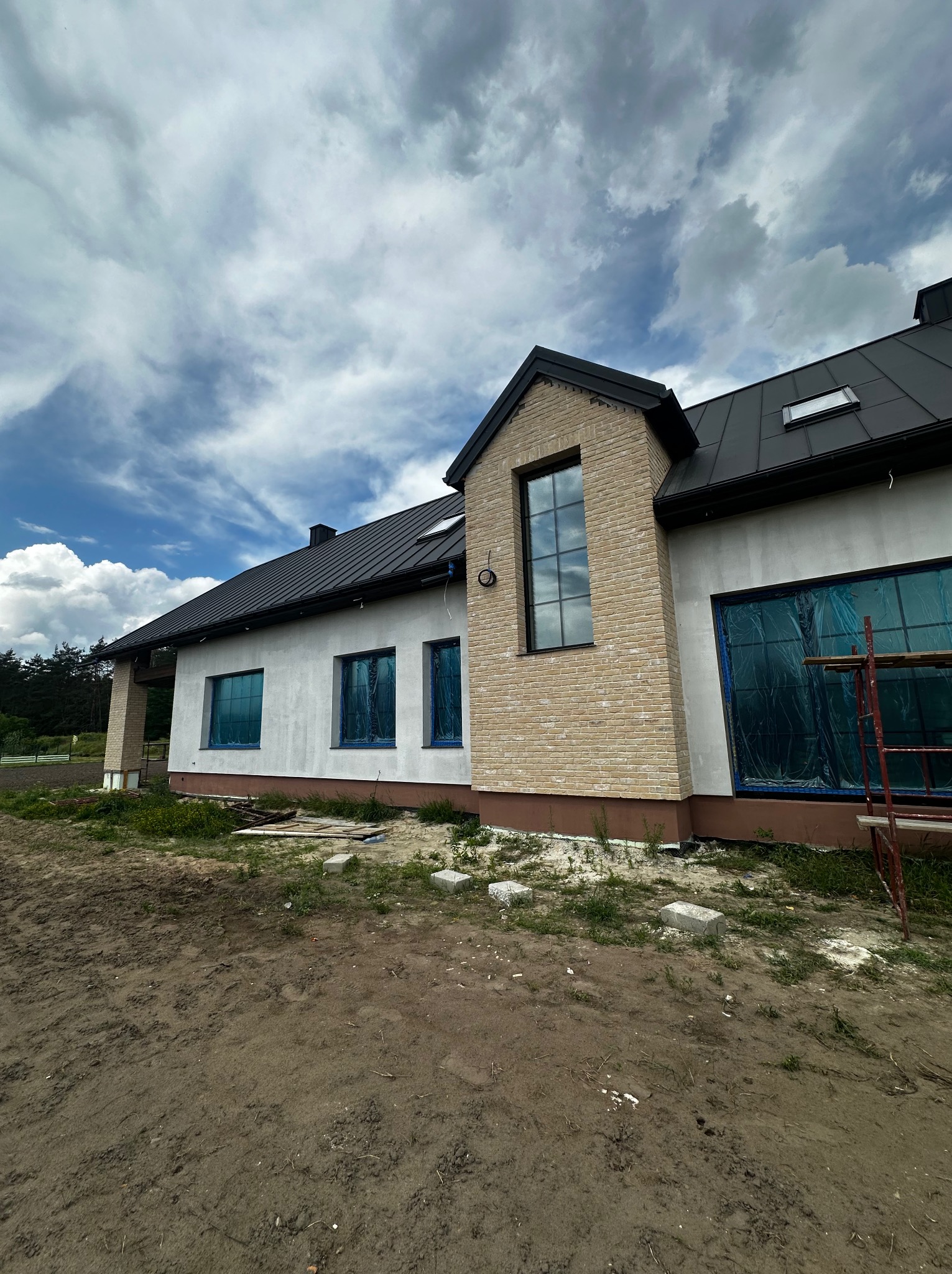 Elewacja domu jednorodzinnego w trakcie wykończenia, z oknami zabezpieczonymi folią i fragmentem rusztowania. Widoczne detale wykończenia z cegły klinkierowej.