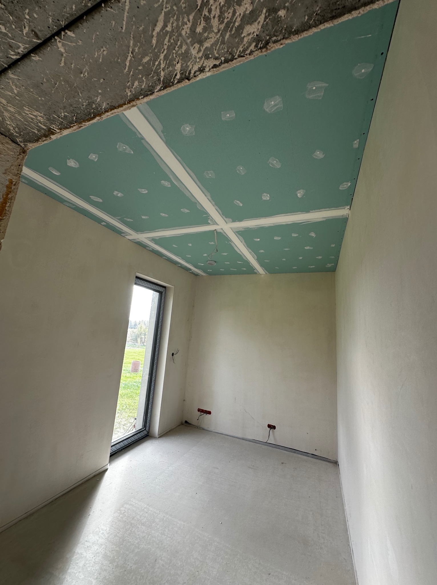 Surowe wnętrze z oknem: ściany po szpachlowaniu, sufit z płyt kartonowo-gipsowych przed dalszym wykończeniem. Widoczne punkty elektryczne.