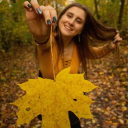 BodziakZaLustrem - Uśmiechnięta brunetka w jesiennym lesie trzyma przed sobą duży, żółty liść klonu, eksponując manicure z brokatem. Tło stanowią drzewa i opadłe liście.