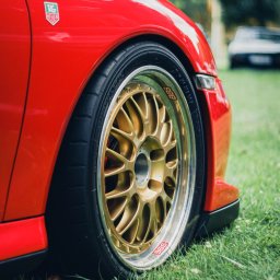 BodziakZaLustrem - Czerwony samochód sportowy z felgami BBS w kolorze złotym, na tle zielonej trawy. Widoczny znaczek TAG Heuer. Ujęcie z bliska, podkreślające detale.