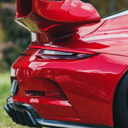 BodziakZaLustrem - Czerwony sportowy samochód z dużym spojlerem, widok z tyłu, na trawie. Detale karoserii, światła i wydech z włókna węglowego. Błyszcząca powierzchnia odbija światło.