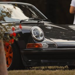 BodziakZaLustrem - Czarny, klasyczny samochód Porsche 911 z pomarańczowymi felgami, sfotografowany z niskiej perspektywy na trawie, z rozmyciem tła i elementami roślin w kadrze.