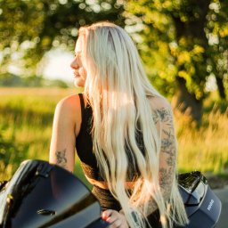 BodziakZaLustrem - Blondwłosa kobieta z tatuażami siedzi na motocyklu w Wierzeja, spoglądając w dal. Ujęcie w ciepłym świetle, z rozmytym tłem zieleni.