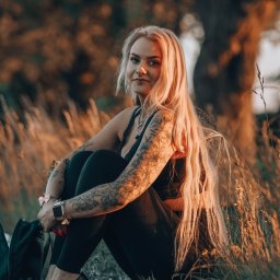 BodziakZaLustrem - Portret kobiety z tatuażami, siedzącej w trawie o zachodzie słońca. Blond włosy, czarny strój, wysokie obcasy. Ujęcie z dołu, ciepłe światło.