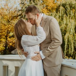 BodziakZaLustrem - Jesienne ujęcie całującej się pary młodej na tle drzew z żółtymi liśćmi. Panna młoda w okularach i sukni z koronką, pan młody w beżowym garniturze.