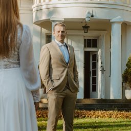 BodziakZaLustrem - Elegancki pan młody w beżowym garniturze, z rękami w kieszeniach, patrzy w obiektyw. W tle biały budynek z kolumnami i częściowo widoczna panna młoda w białej sukni.
