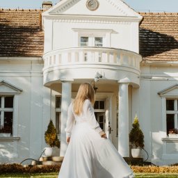BodziakZaLustrem - Panna młoda w białej sukni, tyłem, na tle białego dworku z balkonem i zegarem. Ujęcie w słoneczny dzień, na trawniku.