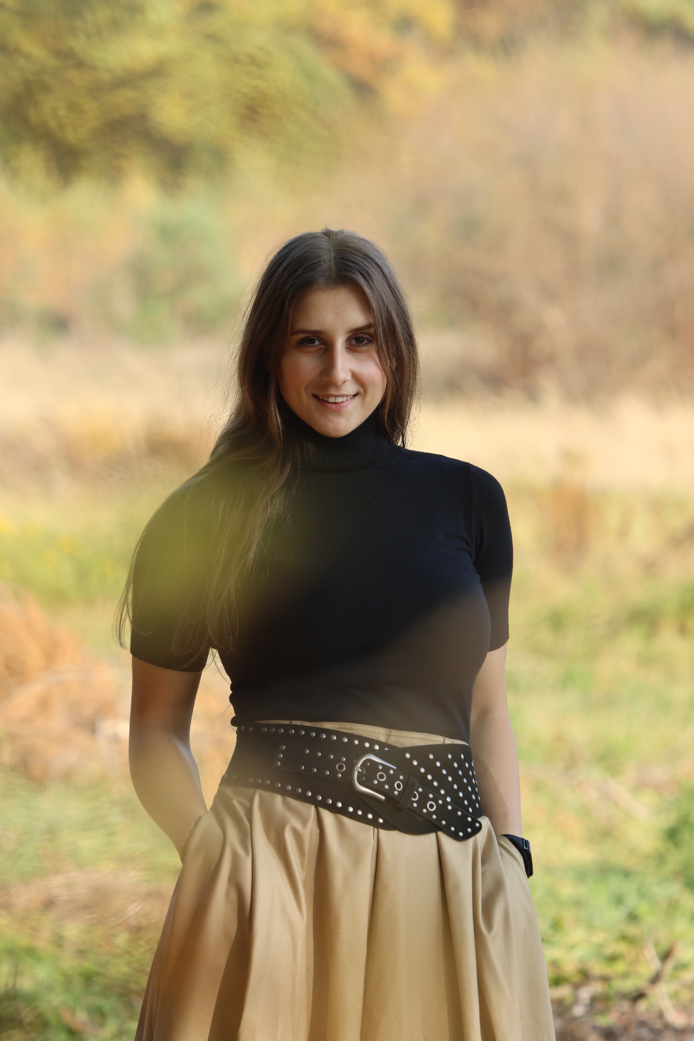 Portret uśmiechniętej brunetki w czarnym golfie i beżowej spódnicy z szerokim paskiem na tle jesiennego krajobrazu. Naturalne światło, delikatne rozmycie tła.