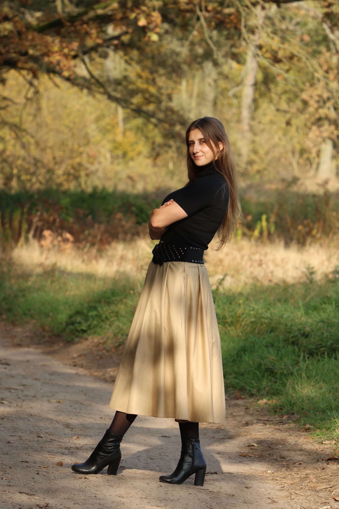 Portret kobiety w czarnym golfie i beżowej spódnicy na tle jesiennego lasu. Ujęcie z dołu, podkreślające sylwetkę i naturalne światło.