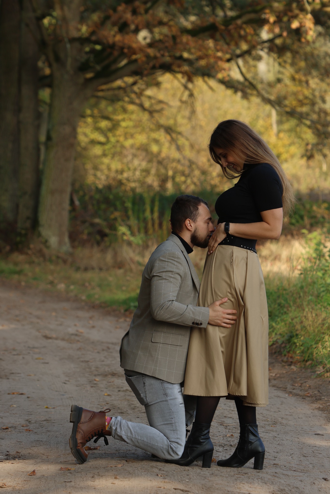 Mężczyzna klęczy, całując brzuch kobiety w beżowej spódnicy na leśnej ścieżce. Sesja ciążowa w plenerze, jesienne tło z drzewami.