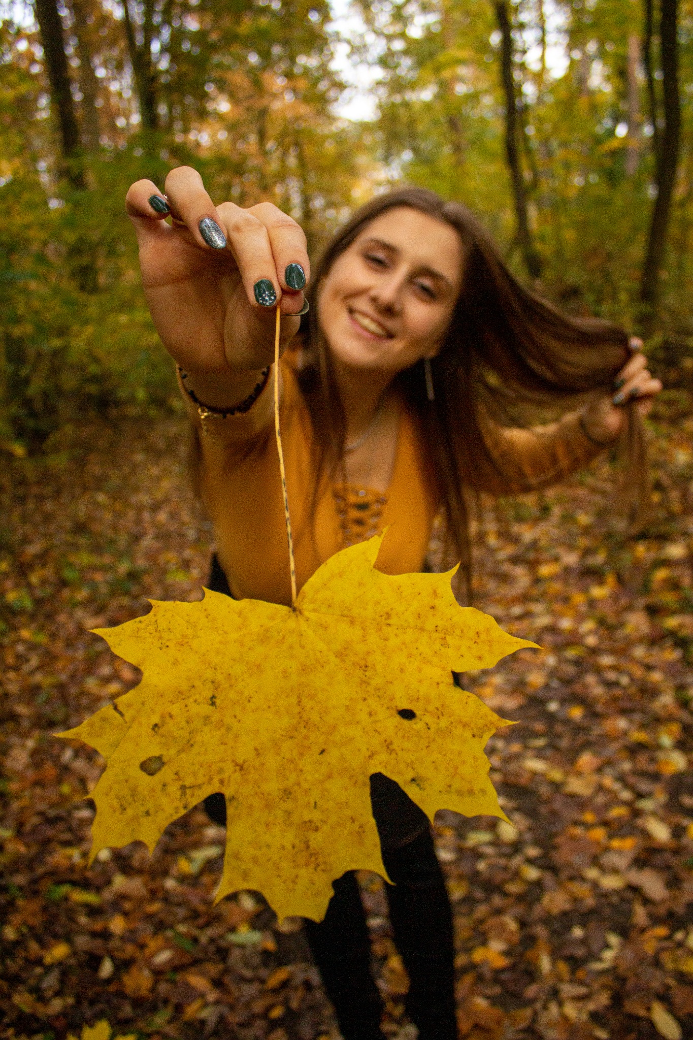 Uśmiechnięta brunetka w jesiennym lesie trzyma przed sobą duży, żółty liść klonu, eksponując manicure z brokatem. Tło stanowią drzewa i opadłe liście.