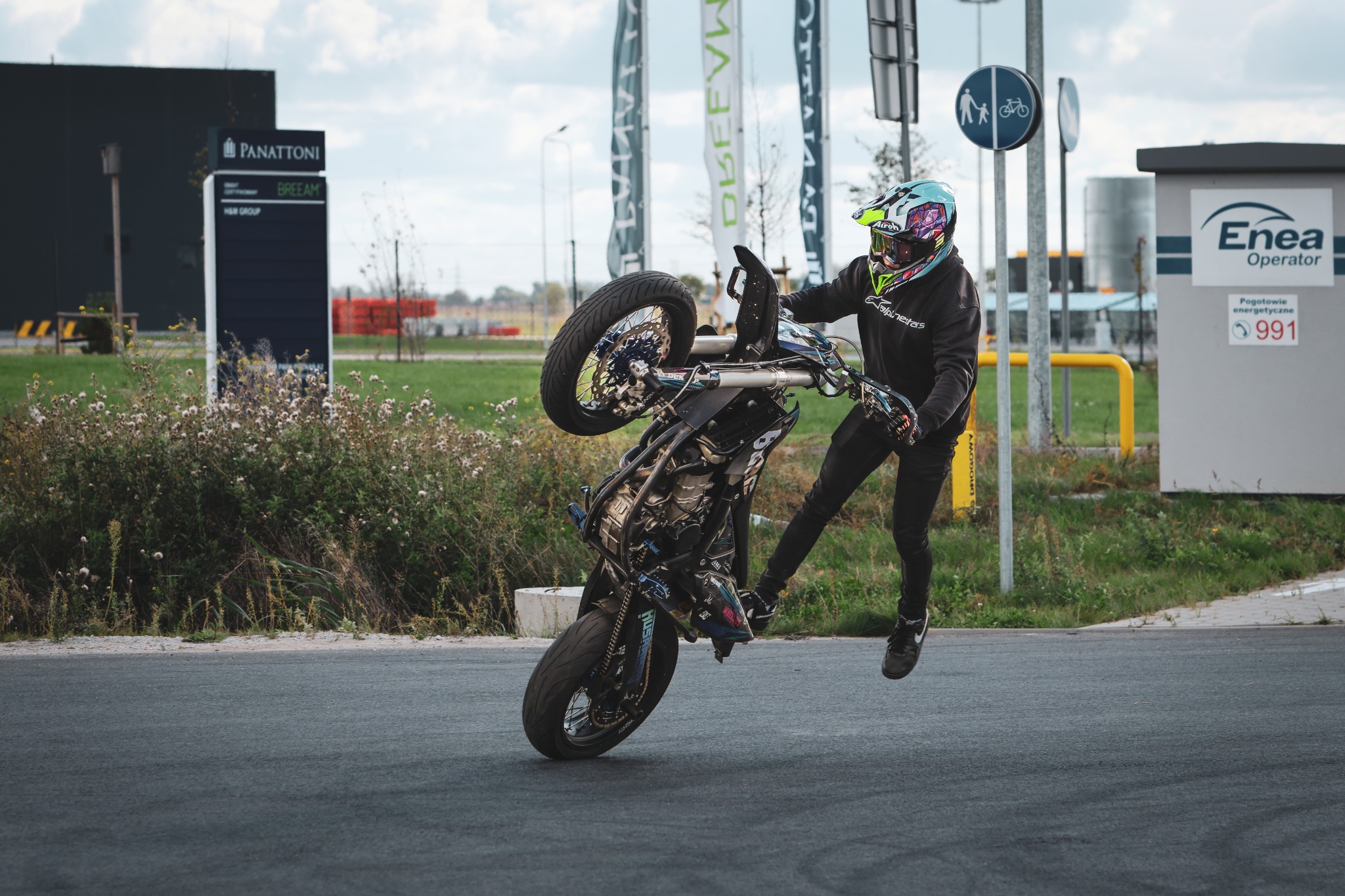 Mężczyzna w kasku i kombinezonie wykonuje trik na motocyklu, unosząc go na jednym kole. W tle budynki i flagi, dynamiczne ujęcie.