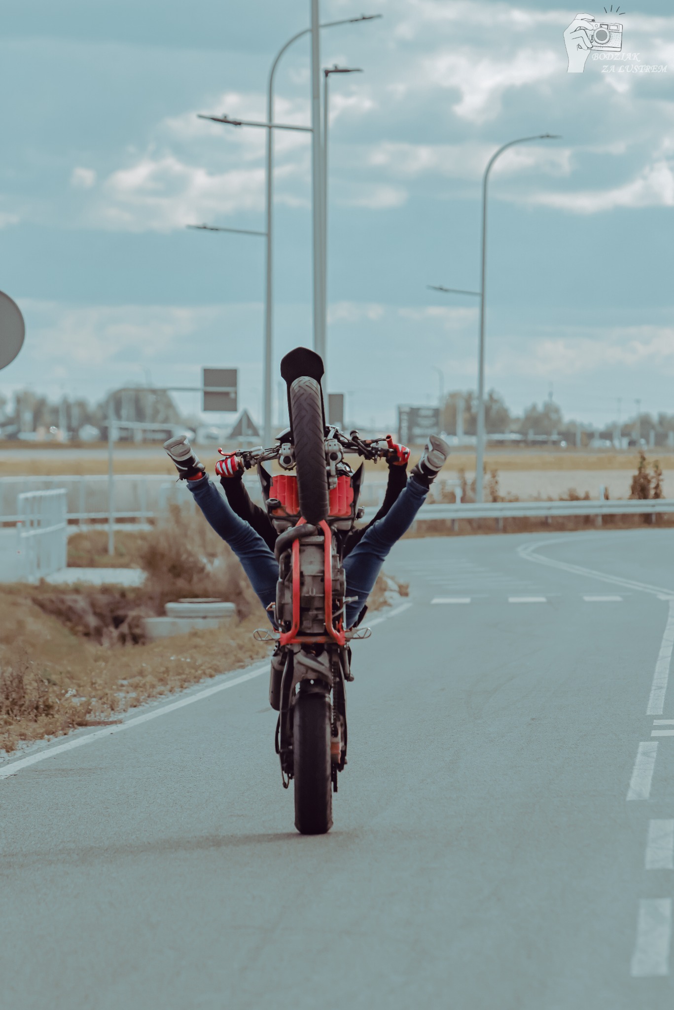Motocykl jadący na jednym kole, z nogami kierowcy uniesionymi do góry, na tle drogi i nieba. Ujęcie z tyłu, podkreślające akrobacje motocyklowe.