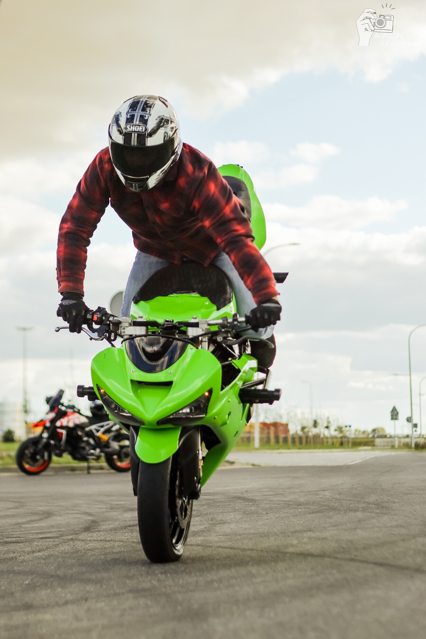 Kaskader na zielonym motocyklu sportowym, wykonujący wheelie na asfalcie. W tle drugi motocykl i pochmurne niebo. Ujęcie z dołu, podkreślające dynamikę.