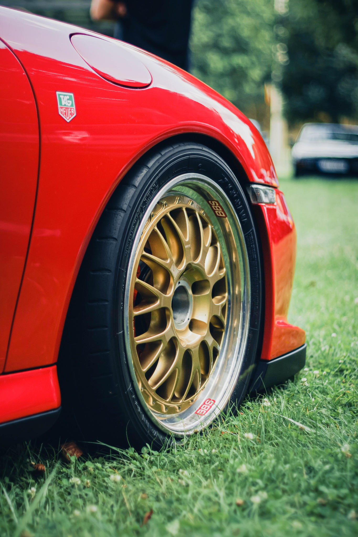 Czerwony samochód sportowy z felgami BBS w kolorze złotym, na tle zielonej trawy. Widoczny znaczek TAG Heuer. Ujęcie z bliska, podkreślające detale.