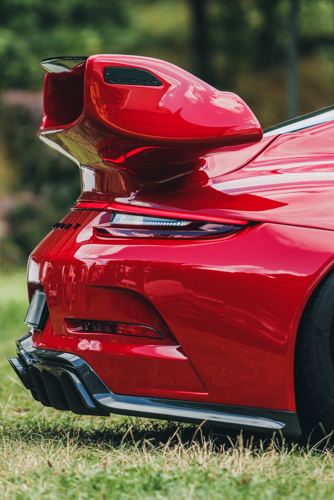 Czerwony sportowy samochód z dużym spojlerem, widok z tyłu, na trawie. Detale karoserii, światła i wydech z włókna węglowego. Błyszcząca powierzchnia odbija światło.