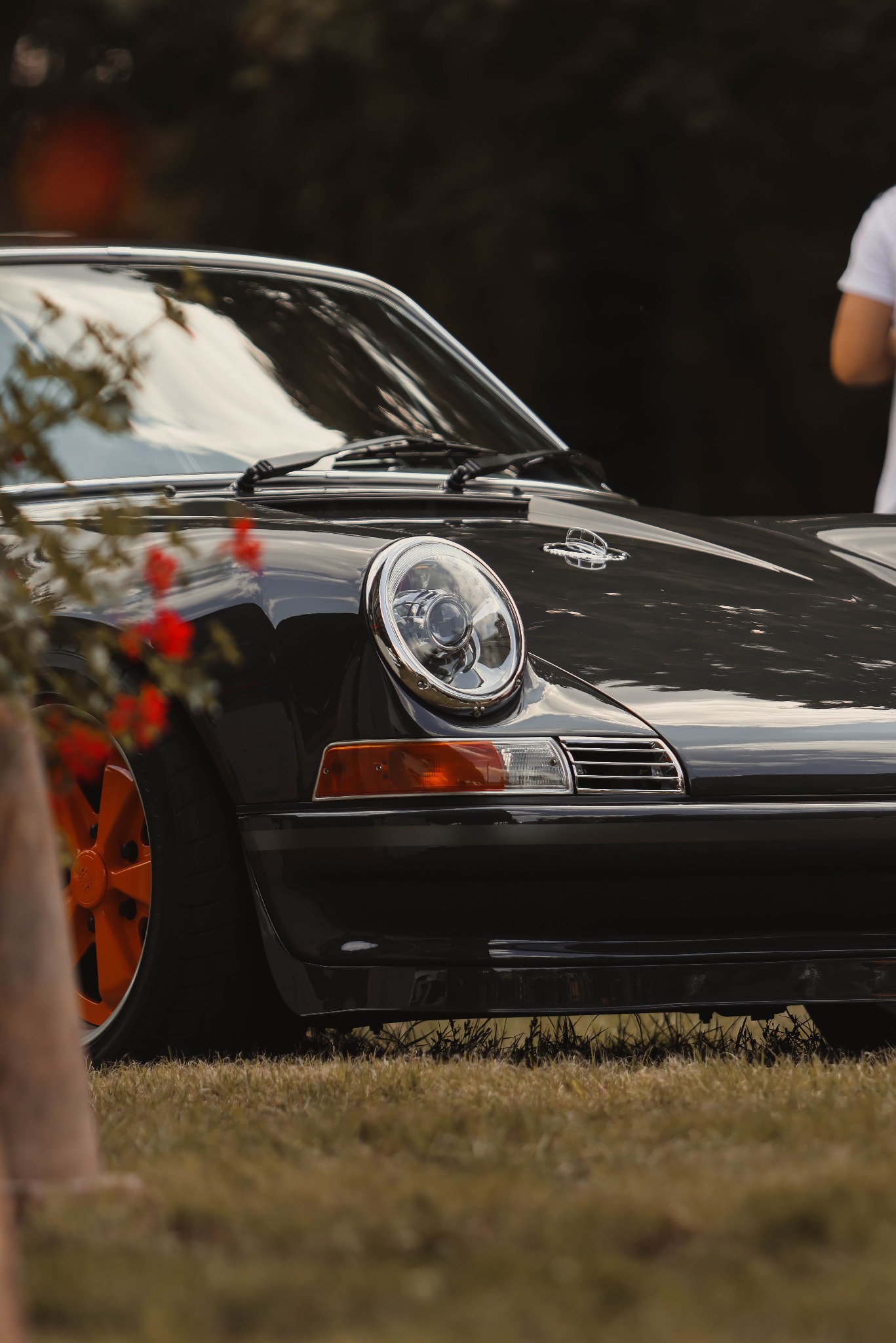 Czarny, klasyczny samochód Porsche 911 z pomarańczowymi felgami, sfotografowany z niskiej perspektywy na trawie, z rozmyciem tła i elementami roślin w kadrze.