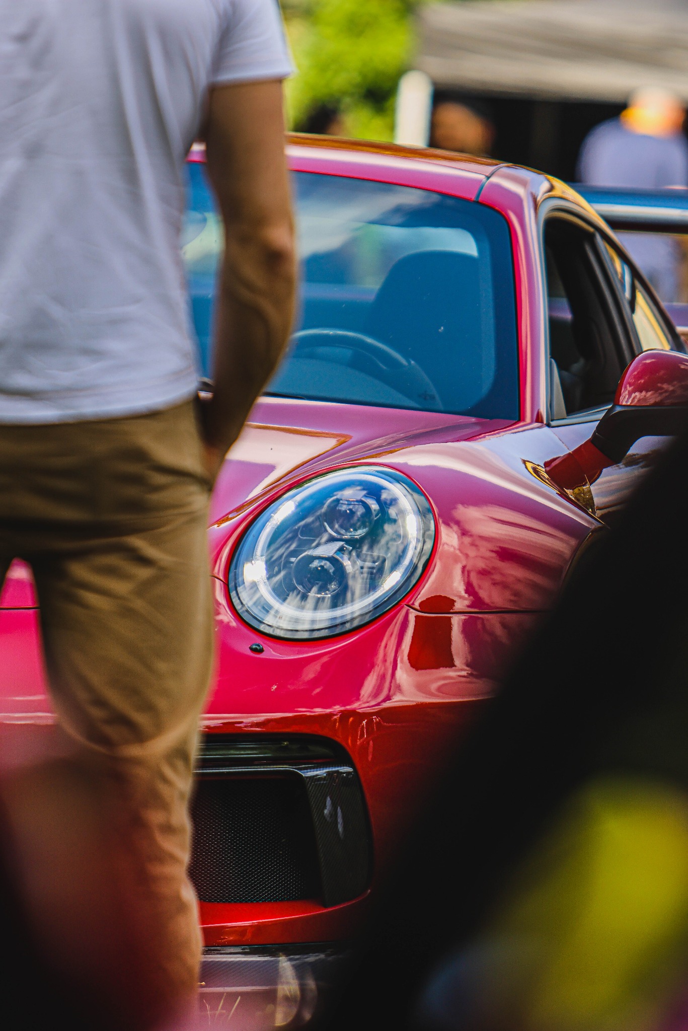 Czerwony sportowy samochód z widocznym reflektorem i fragmentem sylwetki osoby w tle, uchwycony z bliska, z perspektywy podkreślającej detale karoserii.