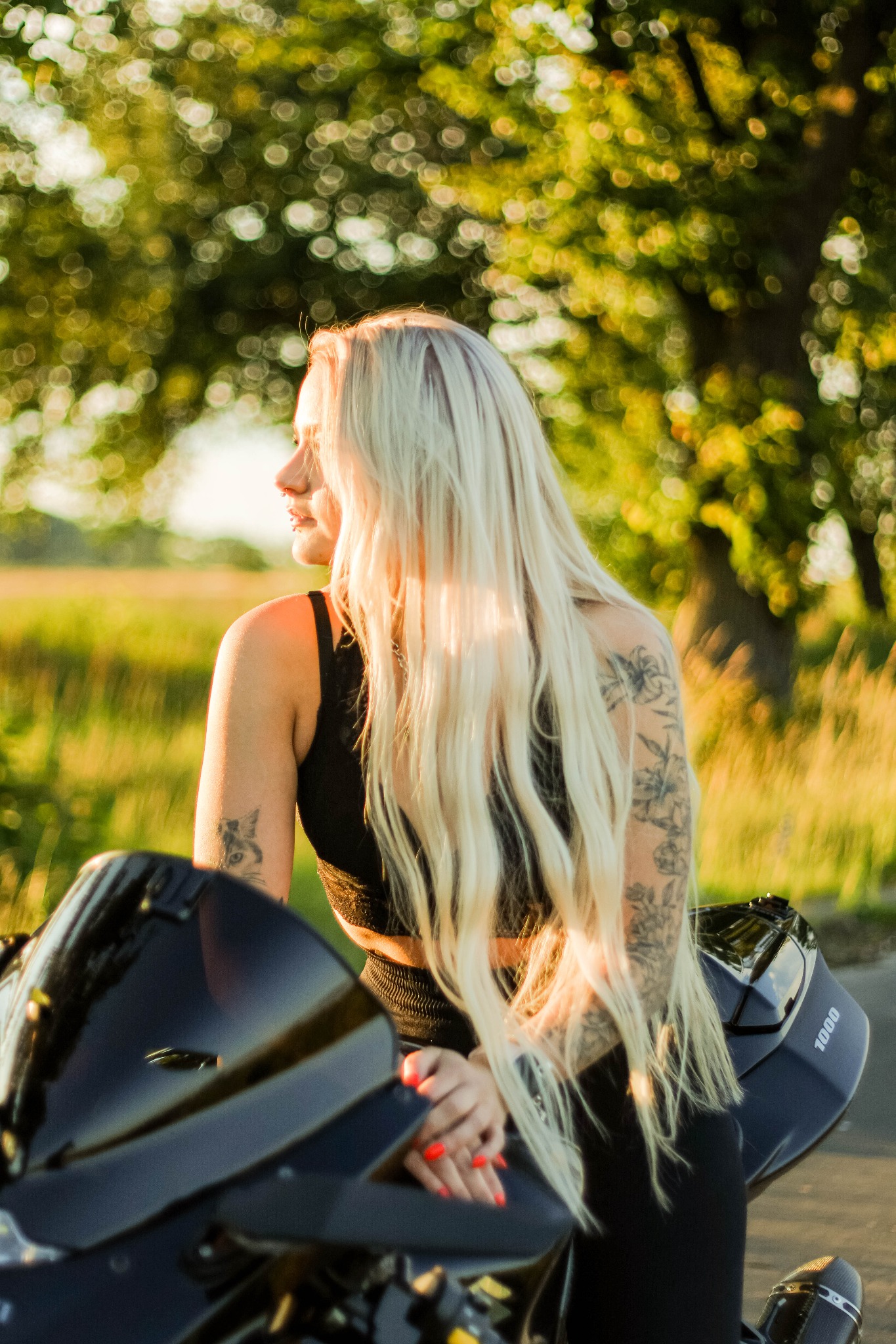 Blondwłosa kobieta z tatuażami siedzi na motocyklu w Wierzeja, spoglądając w dal. Ujęcie w ciepłym świetle, z rozmytym tłem zieleni.