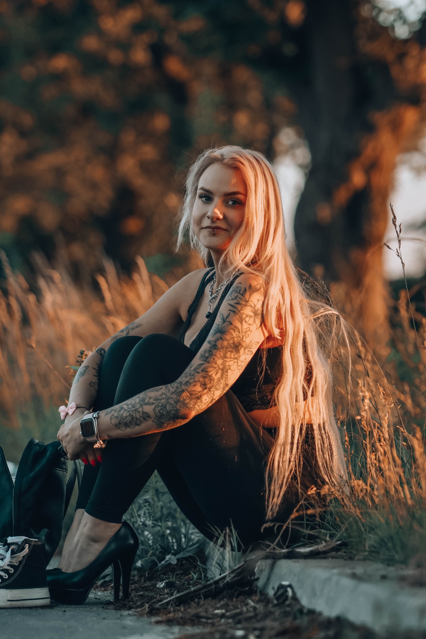 Portret kobiety z tatuażami, siedzącej w trawie o zachodzie słońca. Blond włosy, czarny strój, wysokie obcasy. Ujęcie z dołu, ciepłe światło.