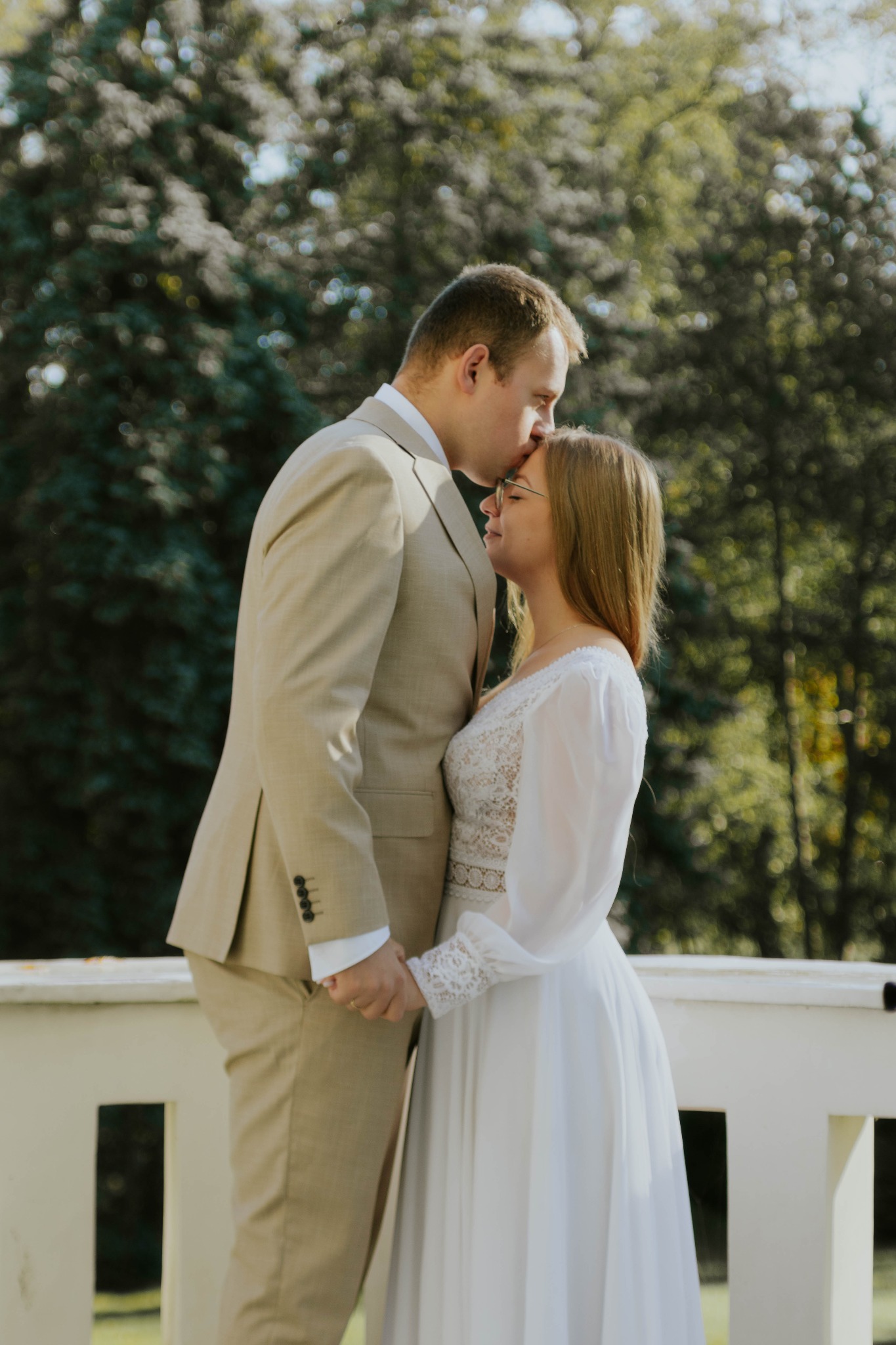 Para młoda w objęciach na tle zieleni. Mężczyzna w beżowym garniturze całuje kobietę w białej sukni w czoło. Delikatne światło podkreśla intymność chwili.