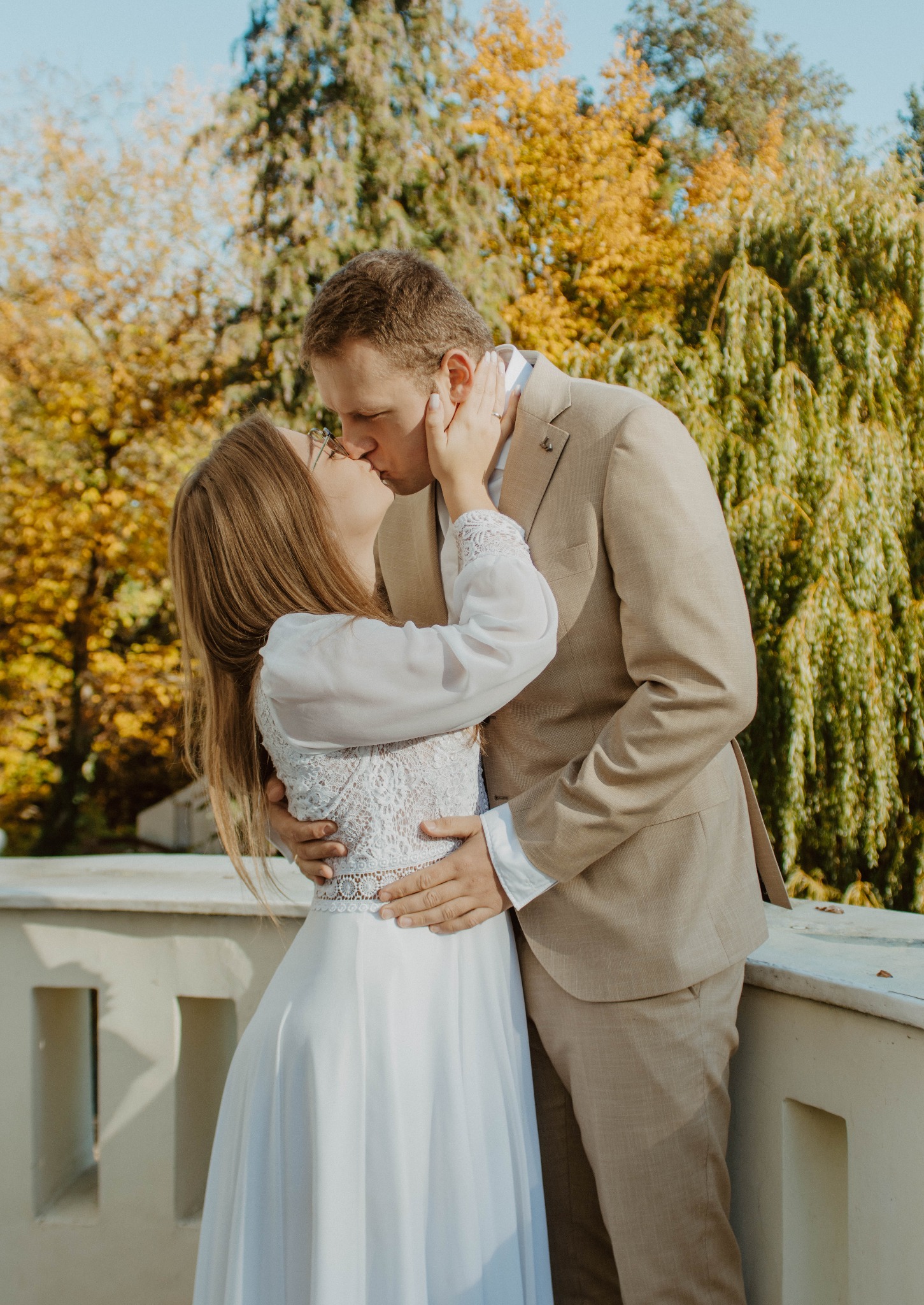 Jesienne ujęcie całującej się pary młodej na tle drzew z żółtymi liśćmi. Panna młoda w okularach i sukni z koronką, pan młody w beżowym garniturze.