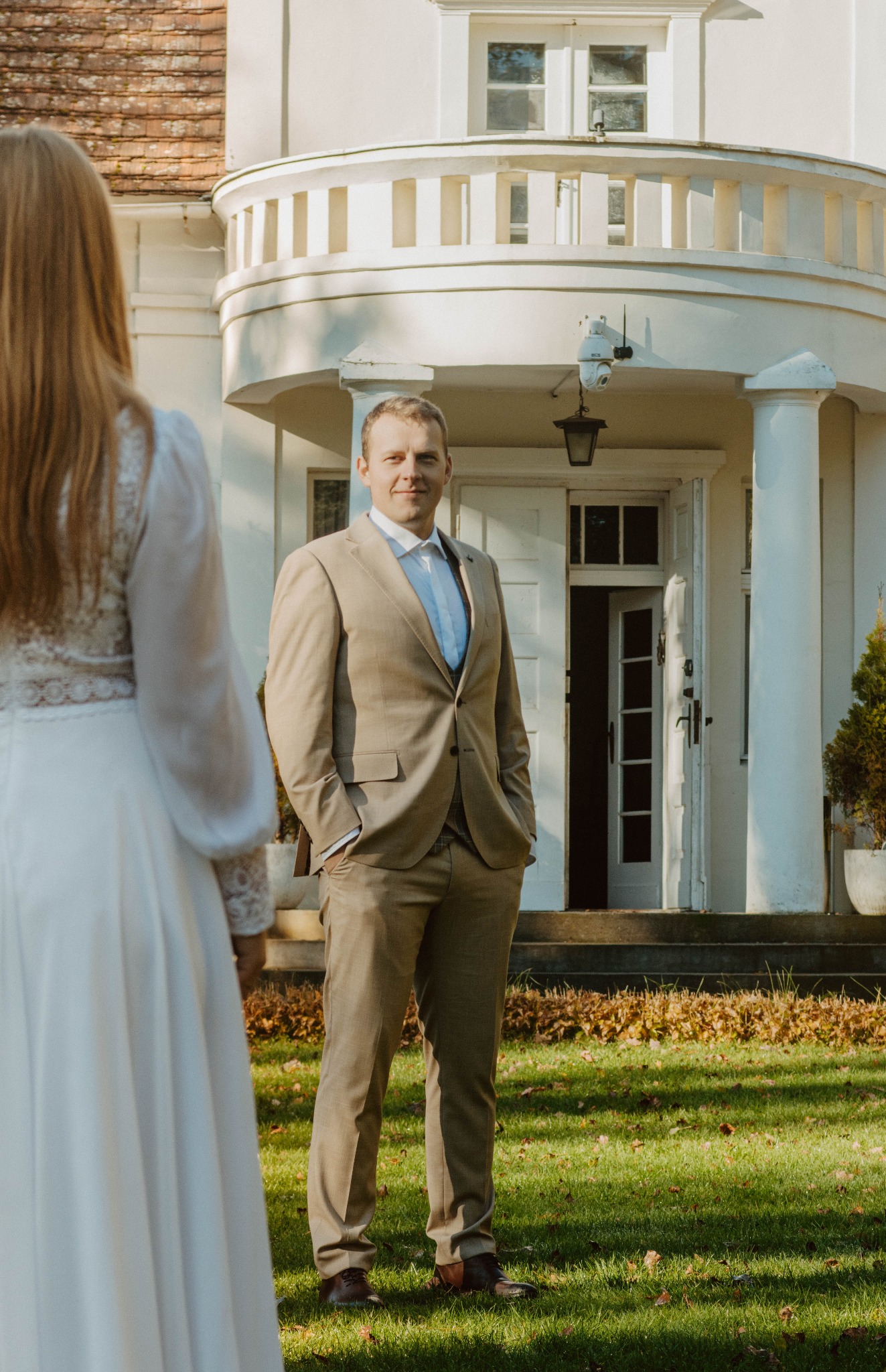 Elegancki pan młody w beżowym garniturze, z rękami w kieszeniach, patrzy w obiektyw. W tle biały budynek z kolumnami i częściowo widoczna panna młoda w białej sukni.