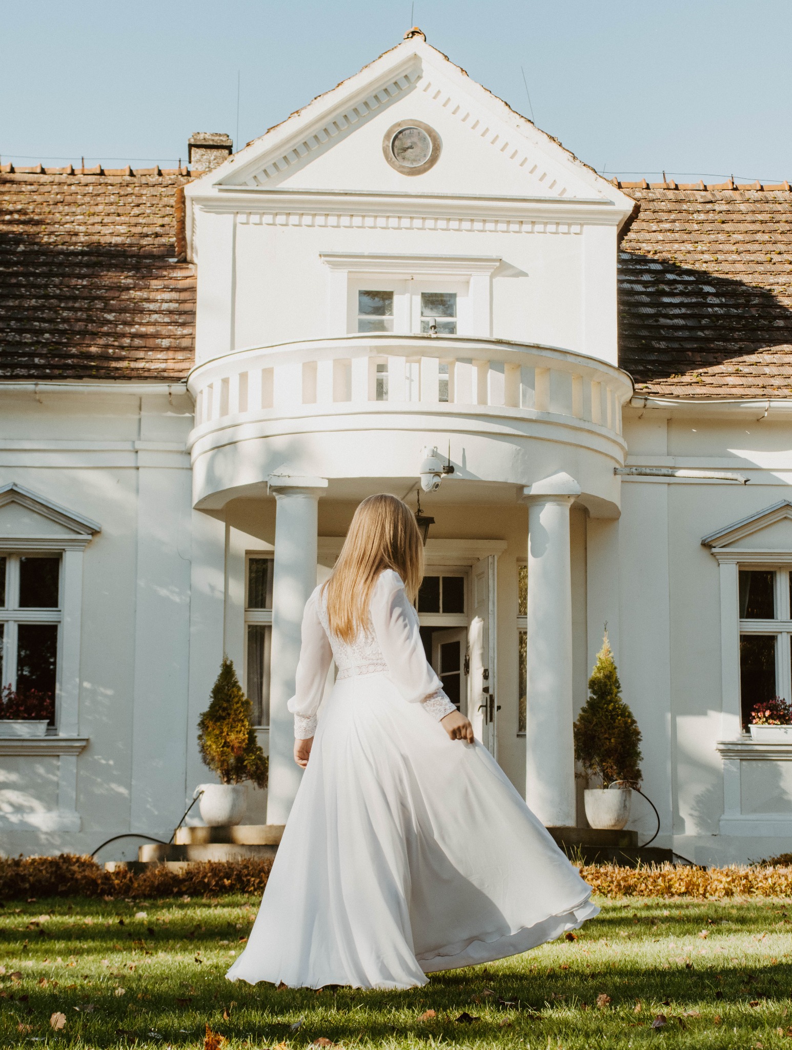Panna młoda w białej sukni, tyłem, na tle białego dworku z balkonem i zegarem. Ujęcie w słoneczny dzień, na trawniku.