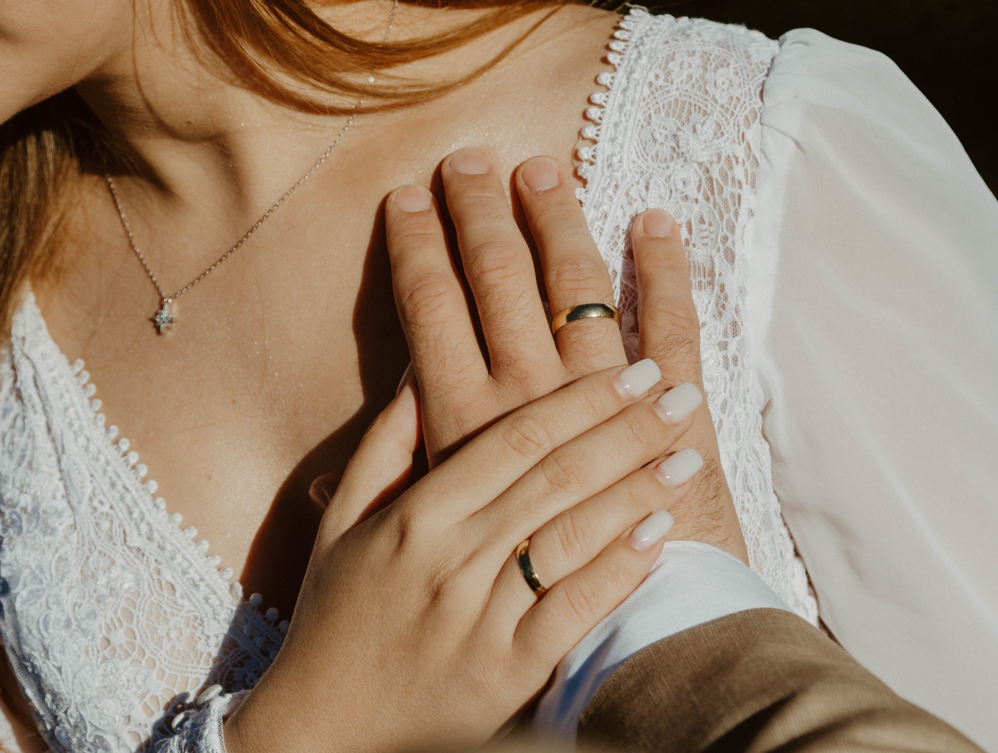 Zbliżenie dłoni pary młodej z złotymi obrączkami na tle koronkowej sukni ślubnej i brązowego garnituru. Delikatne światło podkreśla detale.