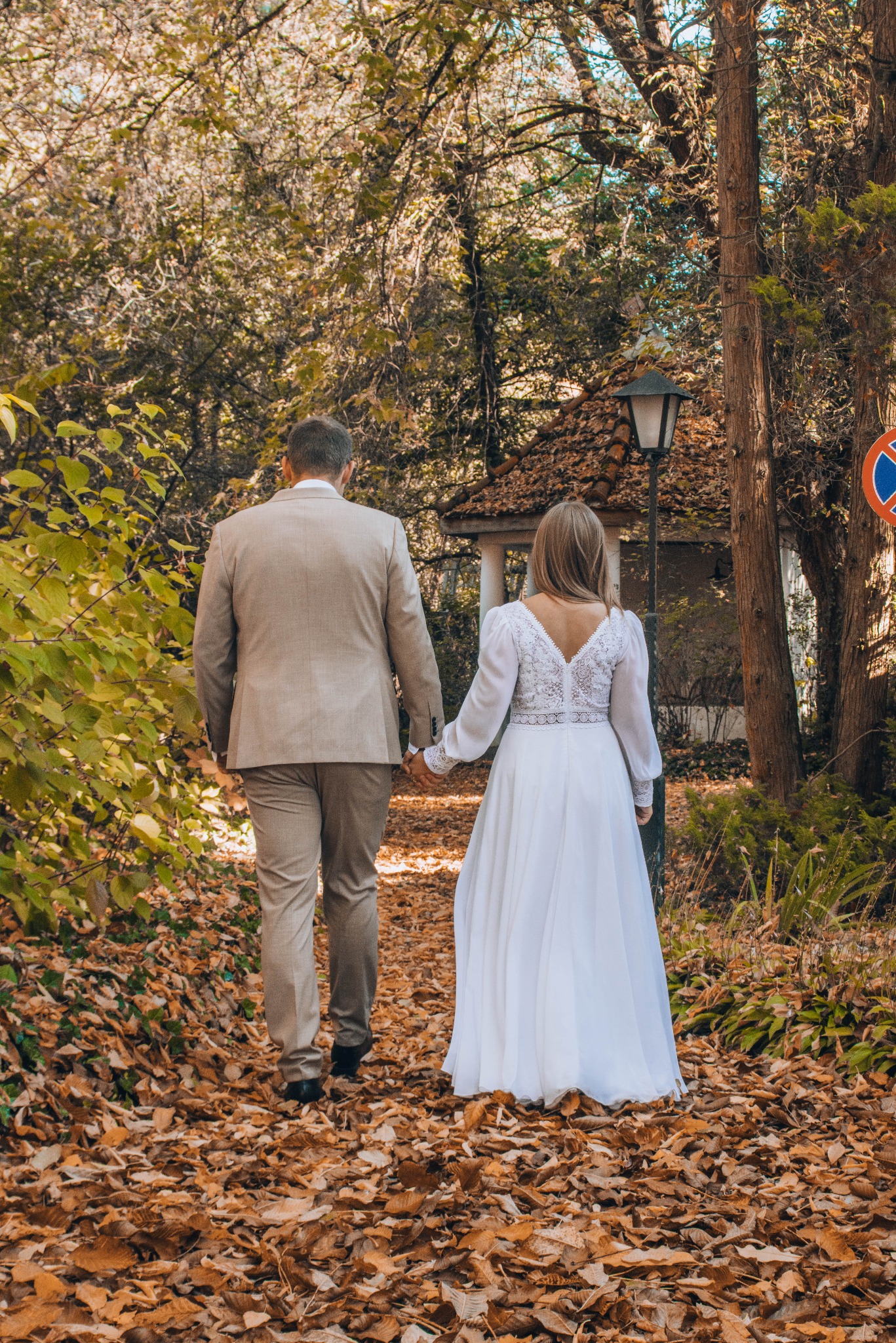 Para młoda, trzymając się za ręce, spaceruje aleją usłaną jesiennymi liśćmi. Panna młoda w białej sukni z koronkowymi plecami, pan młody w beżowym garniturze.