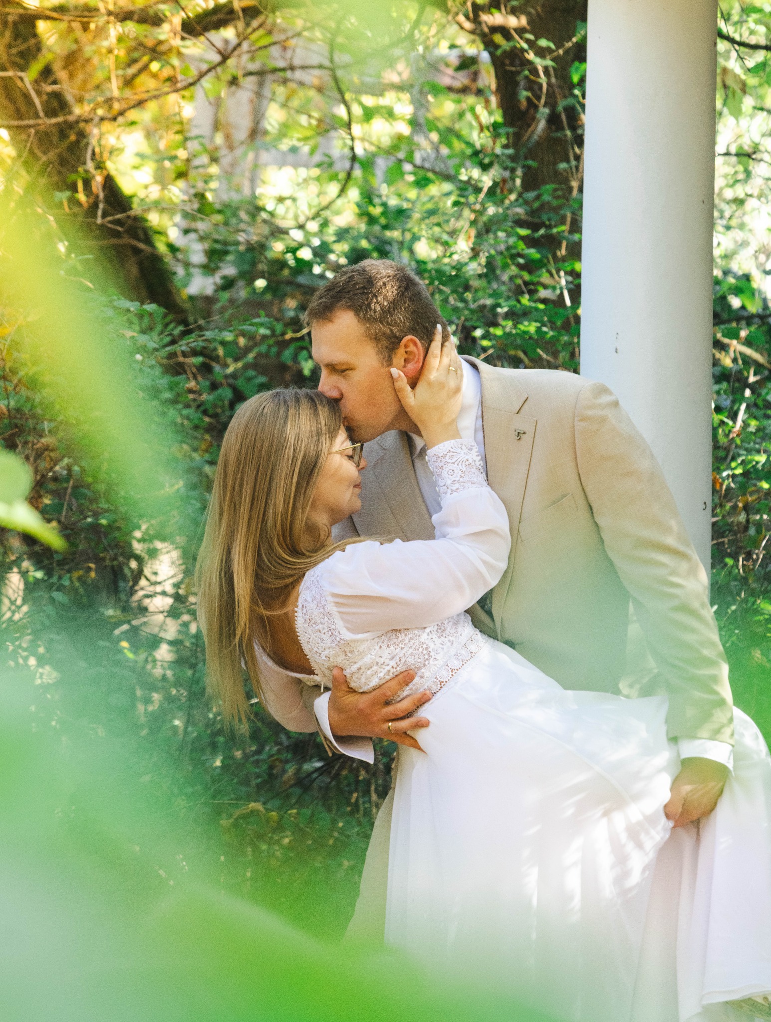 Para młoda w objęciach w plenerze: on całuje ją w czoło, ona trzyma jego twarz. Romantyczna sesja ślubna w otoczeniu zieleni i białej kolumny.